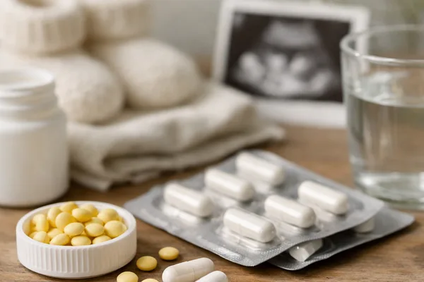 Weiße Kapseln im Blister, gelbe Tabletten, Wasserglas, Babyschuhe und Ultraschallbild auf Holztisch