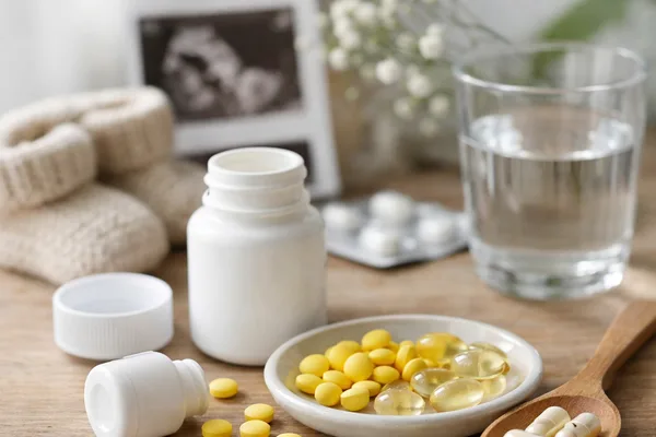 Gelbe Tabletten und Ölkapseln in Schale, weiße Dosen, Wasserglas, Ultraschallbild und Babyschuhe